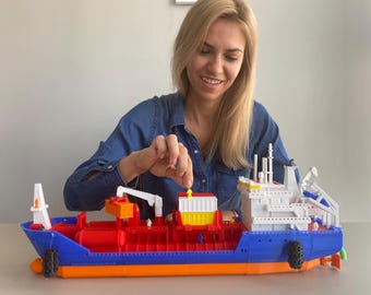 Könnte beinhalten: Eine Frau spielt mit einem großen, blauen, roten und orangenen Spielzeugschiff. Das Schiff hat eine weiße Kabine und einen gelben Frachtcontainer. Die Frau hält einen kleinen gelben Container in der Hand.