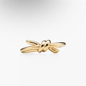 May include: A polished gold ring featuring a continuous band that loops and crosses over itself in the center to form a knot design. The metal surface is smooth and reflective, showcasing a warm yellow tone throughout the entire circular structure.