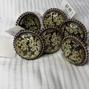 May include: A collection of six round cabinet knobs featuring a dark brown background with a light yellow floral and leaf pattern. Each knob has a smooth, domed glass surface encased in a metallic frame with a beaded edge detail.