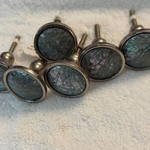 May include: A set of five round cabinet knobs featuring a dark, iridescent, cracked shell inlay set within a metallic silver-toned frame. Each knob has a short, cylindrical stem and a circular face with a mosaic-like pattern of dark gray and blue.