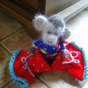 May include: A gray knitted toy mouse wearing a blue, yellow, and red knitted dress with a red knitted skirt. The skirt has a blue trim and is decorated with white polka dots.