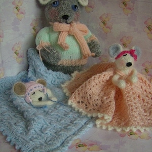 May include: Two knitted mouse toys, one in a light blue sweater and the other in a pink dress. The mouse in the sweater is lying down in a blue knitted blanket, while the mouse in the dress is standing up. Both mice have gray fur and black eyes.