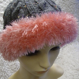 May include: A gray knitted hat with a pink fluffy trim. The hat is on a white fuzzy surface.
