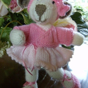 May include: A knitted pink and white ballerina bear with a pink bow. The bear is wearing a pink dress and pink shoes.