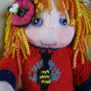 May include: A knitted doll with long, curly, yellow and orange hair. The doll is wearing a red sweater with a blue trim and a bee design. The doll has a pink flower with a bee on top of her head.