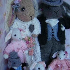 May include: A set of four knitted bunny rabbit dolls dressed in wedding attire. The bride bunny is wearing a white dress with a veil and a bouquet of flowers. The groom bunny is wearing a grey suit with a black top hat and a white bow tie. The two smaller bunnies are wearing pink dresses.