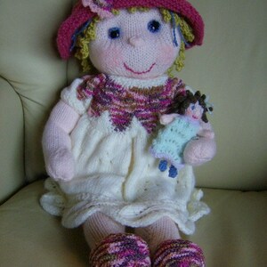May include: A knitted doll with pink and white clothing, a pink and white hat, and a small knitted doll in her arms.
