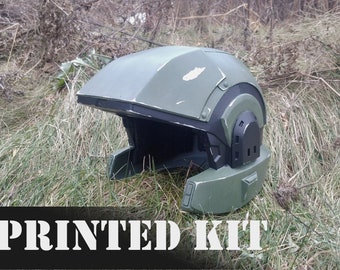 May include: A green and black 3D printed helmet with a weathered look. The helmet is lying on a bed of green grass. The text "PRINTED KIT" is visible at the bottom of the image.