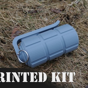 May include: A gray 3D printed replica of a grenade with a ring pull and a handle. The grenade is lying on a bed of dry grass. The text "PRINTED KIT" is visible at the bottom of the image.