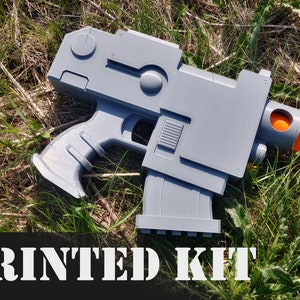 May include: A gray 3D printed gun model with an orange tip. The model is lying on a bed of green grass. The text "PRINTED KIT" is printed in white on a black background.