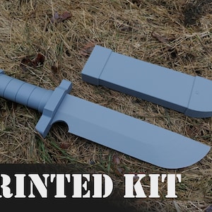 May include: A gray plastic knife and sheath. The knife has a textured handle and a rounded blade. The sheath is a simple rectangular shape. The text "PRINTED KIT" is visible at the bottom of the image.