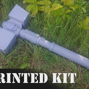 May include: A gray 3D printed hammer with a skull design on the handle. The hammer is lying on a bed of green grass. PRINTED KIT is written in white letters on a black background.