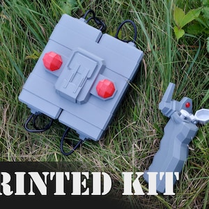 Könnte beinhalten: Ein 3D-gedrucktes Kit für ein Fernsteuerungsgerät. Das Kit enthält einen grauen Controller mit zwei roten Knöpfen und einen grauen Empfänger mit einem roten Knopf. Der Text "PRINTED KIT" ist in Weiß auf schwarzem Hintergrund gedruckt.