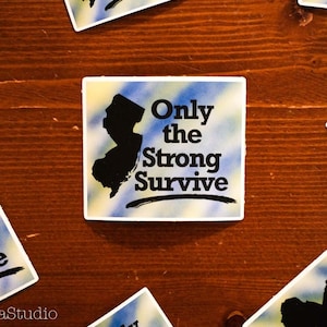 May include: A sticker with a blue and yellow tie-dye background featuring a black silhouette of the state of New Jersey. The sticker has the text "Only the Strong Survive" in black.