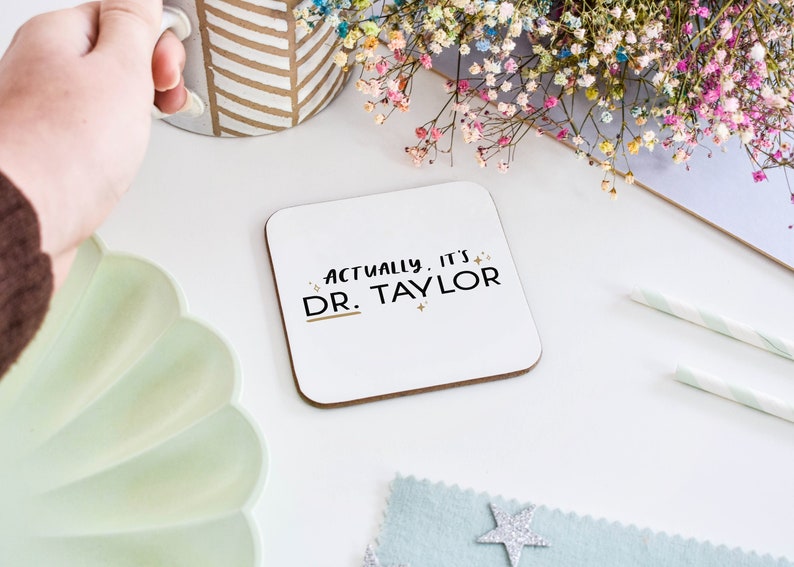 Personalised coaster gift for Doctor graduation gift that reads "Actually it's DR. [chosen name here]" in black typography with a gold-like star pattern