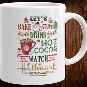 May include: White ceramic mug with a festive design. The mug features the text "Let's Bake Stuff Drink Hot Cocoa and Watch Hallmark Christmas Movies" in red, green, and gold. A red mug of hot cocoa is also depicted.