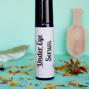 May include: A blue glass roller bottle with a black cap and a white label that reads "Under Eye Serum". The bottle is sitting on a light blue surface with orange flower petals scattered around it.