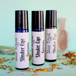 May include: Three glass roller bottles with black caps and white labels. The labels read "Bee Real Under Eye Serum". The bottles are sitting on a light blue surface with dried orange flower petals.