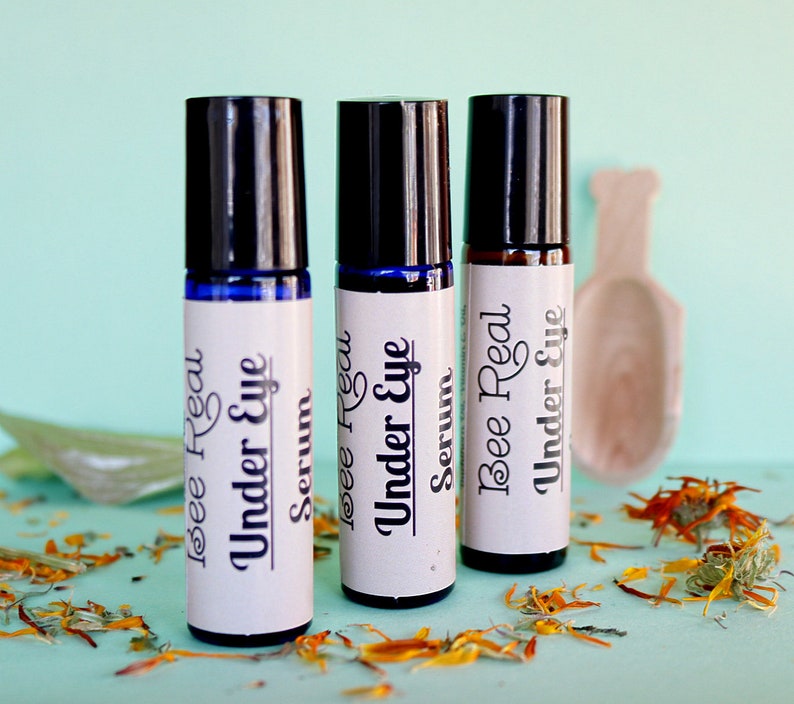 May include: Three glass roller bottles with black caps and white labels. The labels read "Bee Real Under Eye Serum". The bottles are sitting on a light blue surface with dried orange flower petals.