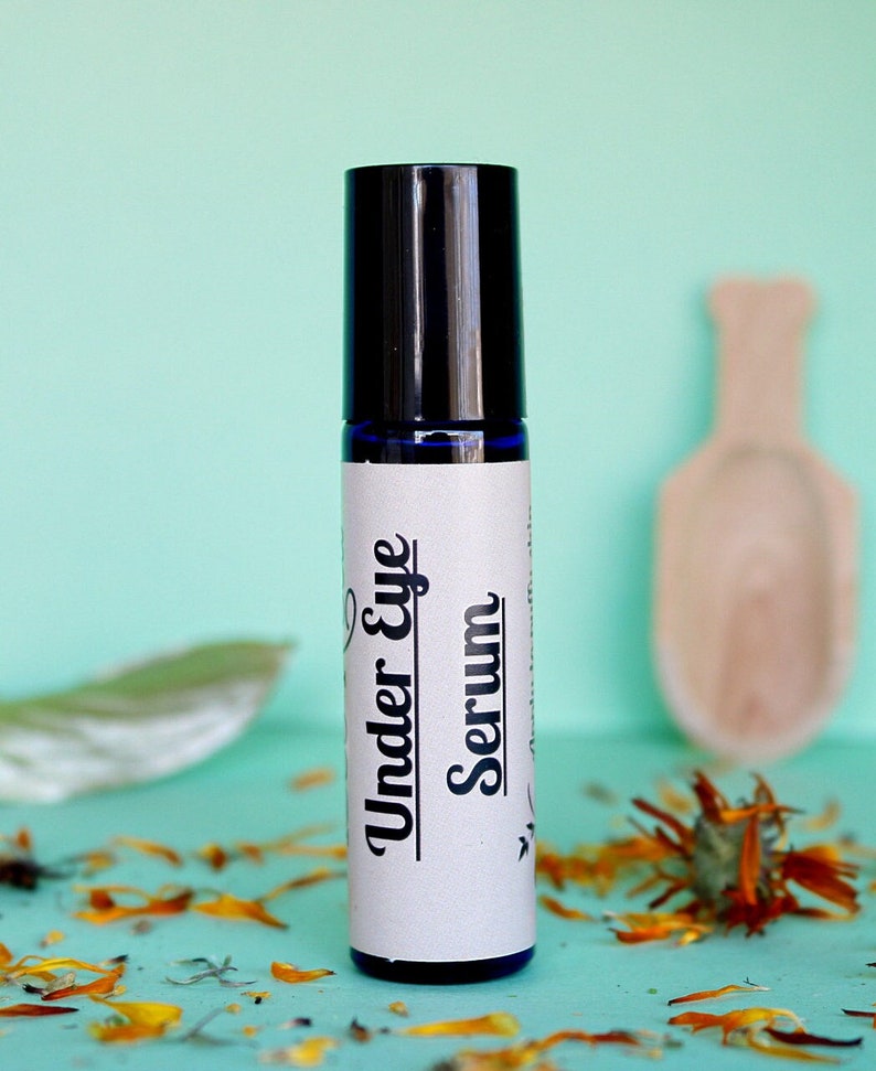 May include: A blue glass roller bottle with a black cap and a white label that reads "Under Eye Serum". The bottle is sitting on a light blue surface with orange flower petals scattered around it.