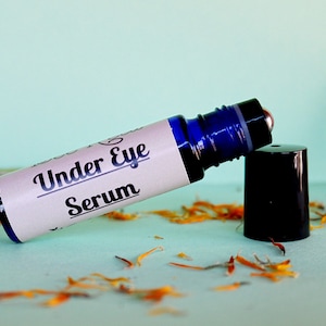 May include: A blue glass roller bottle with a white label that reads "Under Eye Serum". The bottle is lying on a light blue surface with orange flower petals scattered around it.