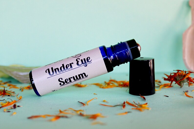 May include: A blue glass roller bottle with a white label that reads "Under Eye Serum". The bottle is lying on a light blue surface with orange flower petals scattered around it.