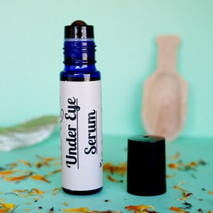 May include: A blue glass roller bottle with a white label that says "Under Eye Serum". The bottle is sitting on a light blue surface with dried flower petals.