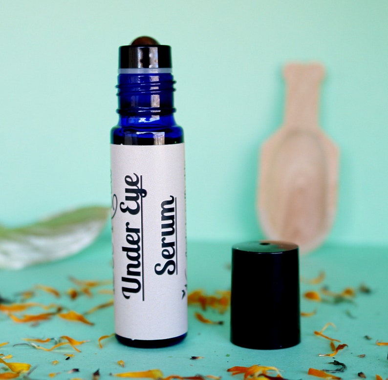 May include: A blue glass roller bottle with a white label that says "Under Eye Serum". The bottle is sitting on a light blue surface with dried flower petals.