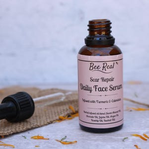 May include: A brown glass bottle with a dropper top and a pink label that reads "Bee Real Scar Repair Daily Face Serum Infused with Turmeric & Calendula Herbal infused oil blend (Sweet Almond Oil, Avocado Oil, Jojoba Oil, Argan Oil, Rosehip Oil, Baobab Oil)"