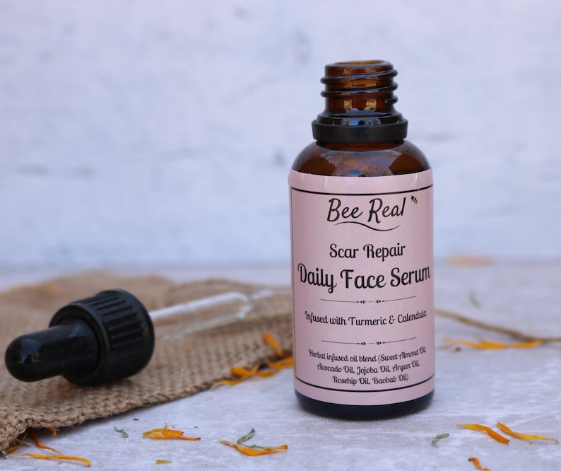 May include: A brown glass bottle with a dropper top and a pink label that reads "Bee Real Scar Repair Daily Face Serum Infused with Turmeric & Calendula Herbal infused oil blend (Sweet Almond Oil, Avocado Oil, Jojoba Oil, Argan Oil, Rosehip Oil, Baobab Oil)"