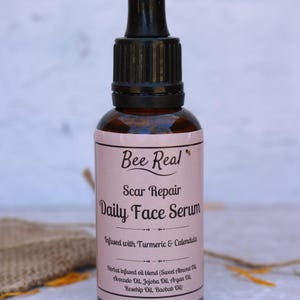 May include: A brown glass bottle with a black dropper top. The label is pink and says "Bee Real Scar Repair Daily Face Serum Infused with Turmeric & Calendula Herbal infused oil blend (Sweet Almond Oil, Avocado Oil, Jojoba Oil, Argan Oil, Rosehip Oil, Baobab Oil)"