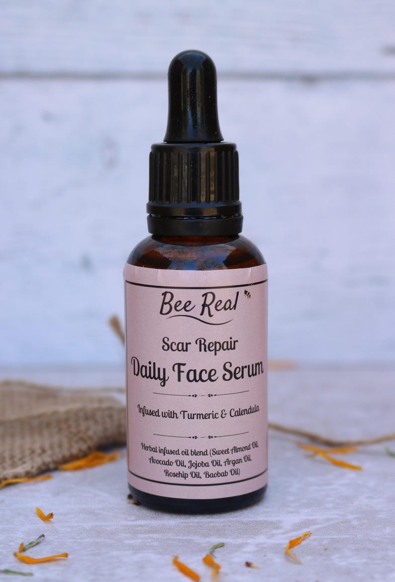 May include: A brown glass bottle with a black dropper top. The label is pink and says "Bee Real Scar Repair Daily Face Serum Infused with Turmeric & Calendula Herbal infused oil blend (Sweet Almond Oil, Avocado Oil, Jojoba Oil, Argan Oil, Rosehip Oil, Baobab Oil)"