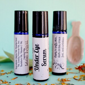 May include: Three blue glass roller bottles with white labels. The labels have black text that reads "Bee Real", "Under Eye Serum", and "Apply to puffy skin, dark circles, crows feet, and wrinkles".