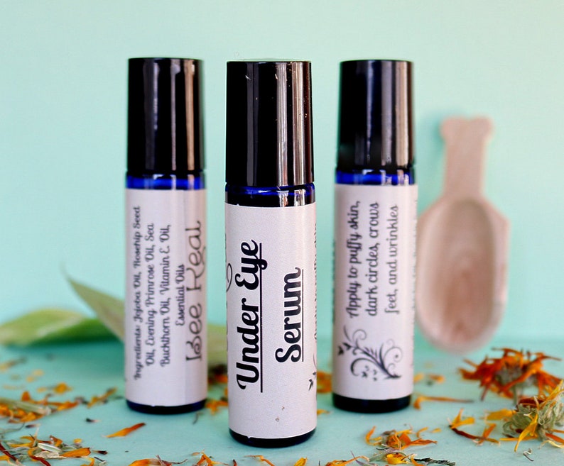 May include: Three blue glass roller bottles with white labels. The labels have black text that reads "Bee Real", "Under Eye Serum", and "Apply to puffy skin, dark circles, crows feet, and wrinkles".