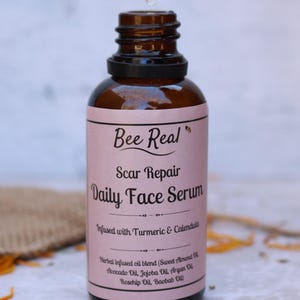May include: A glass bottle with a dropper filled with a clear liquid. The bottle has a pink label with the text "Bee Real Scar Repair Daily Face Serum Infused with Turmeric & Calendula Herbal infused oil blend (Sweet Almond Oil, Avocado Oil, Jojoba Oil, Argan Oil, Rosehip Oil, Baobab Oil)"