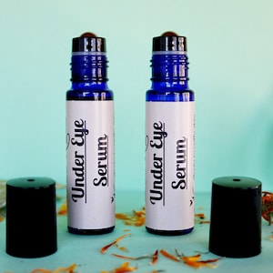 May include: Two blue glass roller bottles with black caps and white labels that say "Under Eye Serum". The bottles are on a light blue surface with orange flower petals.
