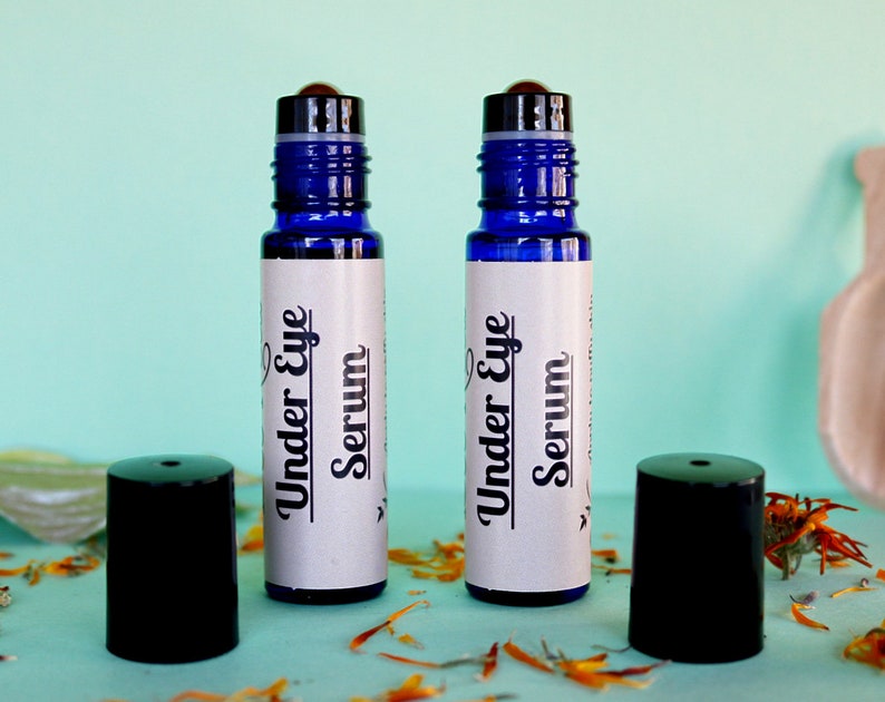 May include: Two blue glass roller bottles with black caps and white labels that say "Under Eye Serum". The bottles are on a light blue surface with orange flower petals.