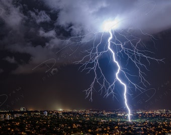 Könnte beinhalten: Ein mächtiger Blitz schlägt durch einen dunklen, stürmischen Himmel über einer Stadt bei Nacht. Der Blitz ist strahlend weiß und beleuchtet die Wolken und die Stadt darunter.