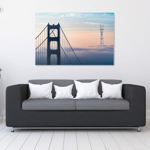 Puede incluir: Un sof&aacute; negro con tres almohadas blancas se encuentra frente a una pared con una impresi&oacute;n enmarcada del Puente Golden Gate en San Francisco. El puente se recorta contra un cielo nublado con una puesta de sol rosa y naranja.