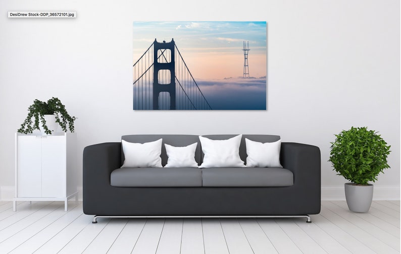 Puede incluir: Un sof&aacute; negro con tres almohadas blancas se encuentra frente a una pared con una impresi&oacute;n enmarcada del Puente Golden Gate en San Francisco. El puente se recorta contra un cielo nublado con una puesta de sol rosa y naranja.