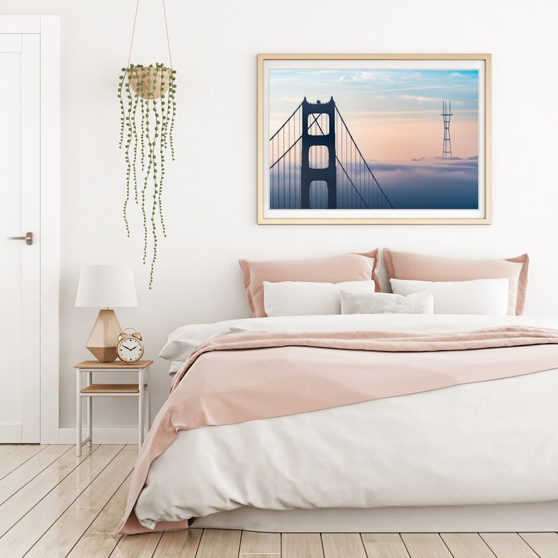 Puede incluir: Un interior de dormitorio con una cama blanca, ropa de cama rosa y una fotograf&iacute;a enmarcada del Puente Golden Gate en San Francisco, California. La fotograf&iacute;a muestra el puente en la distancia con niebla entrando.