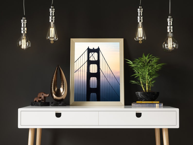 May include: A white desk with two drawers and wooden legs. A framed photograph of the Golden Gate Bridge is on the desk, along with a small wooden elephant figurine, a gold metal abstract sculpture, and a potted green plant.