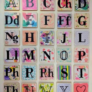 May include: A set of colourful alphabet tiles with various floral and patterned designs. The tiles are arranged in a grid, with each tile featuring a different letter of the alphabet. The tiles are made of wood and have a distressed finish.