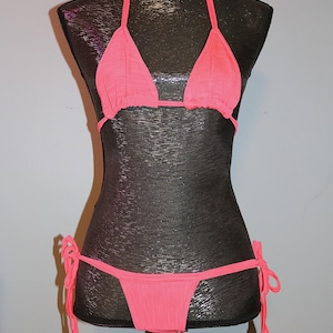 May include: A bright pink bikini set displayed on a mannequin. The top is a triangle style with halter straps, and the bottom is a string bikini. The fabric appears to be a lightweight, textured material. The bikini is tied at the neck and hips.
