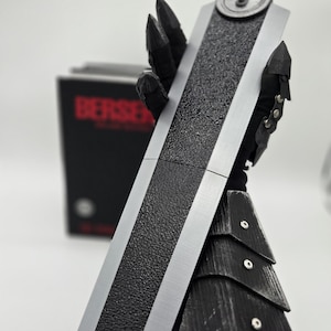 May include: A detailed replica of a large sword with a textured black blade and silver edges. The handle is wrapped in a cream-colored material. The sword is part of a collectible item, with the word "BERSERK" visible on a box in the background.