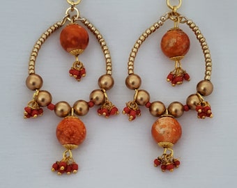 Könnte beinhalten: Ein Paar handgefertigte Ohrringe mit einem goldfarbenen, bogenförmigen Rahmen und kleinen Perlen. Die Ohrringe haben große orange-rote kugelförmige Perlen und kleinere rote Perlencluster.