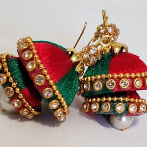 Puede incluir: Un par de pendientes de hilo de seda rojo y verde con detalles dorados y brillantes transparentes. Los pendientes tienen forma de campana y tienen una perla.