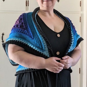 May include: A person wearing a black dress with a blue and purple crocheted shawl. The shawl has a black border and is tied at the front.