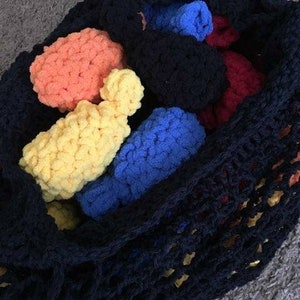May include: A dark navy blue crocheted bag filled with colourful, chunky yarn balls. The yarn balls are in shades of yellow, orange, blue, and red. The bag is on a grey surface.