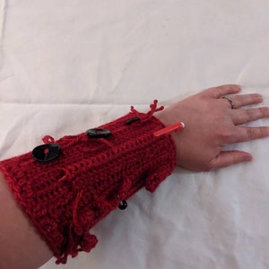 May include: A red crocheted arm cuff with black buttons, beads, and yarn embellishments.